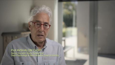 Caspi sits for an interview facing slightly off camera in light grey dress button down. Background is fuzzy and features a room with plenty of natural light.